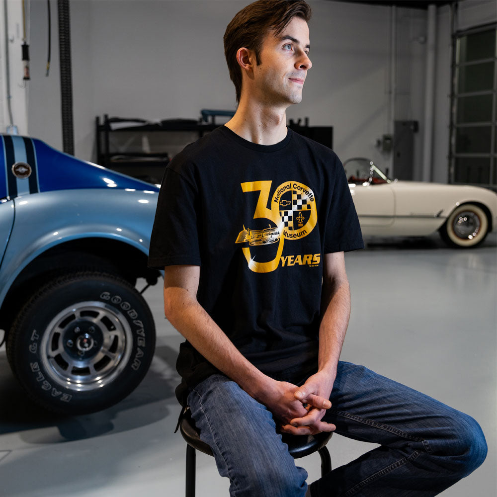 Man sitting on a stool in a garage wearing the National Corvette Museum 30 Years Black Tee