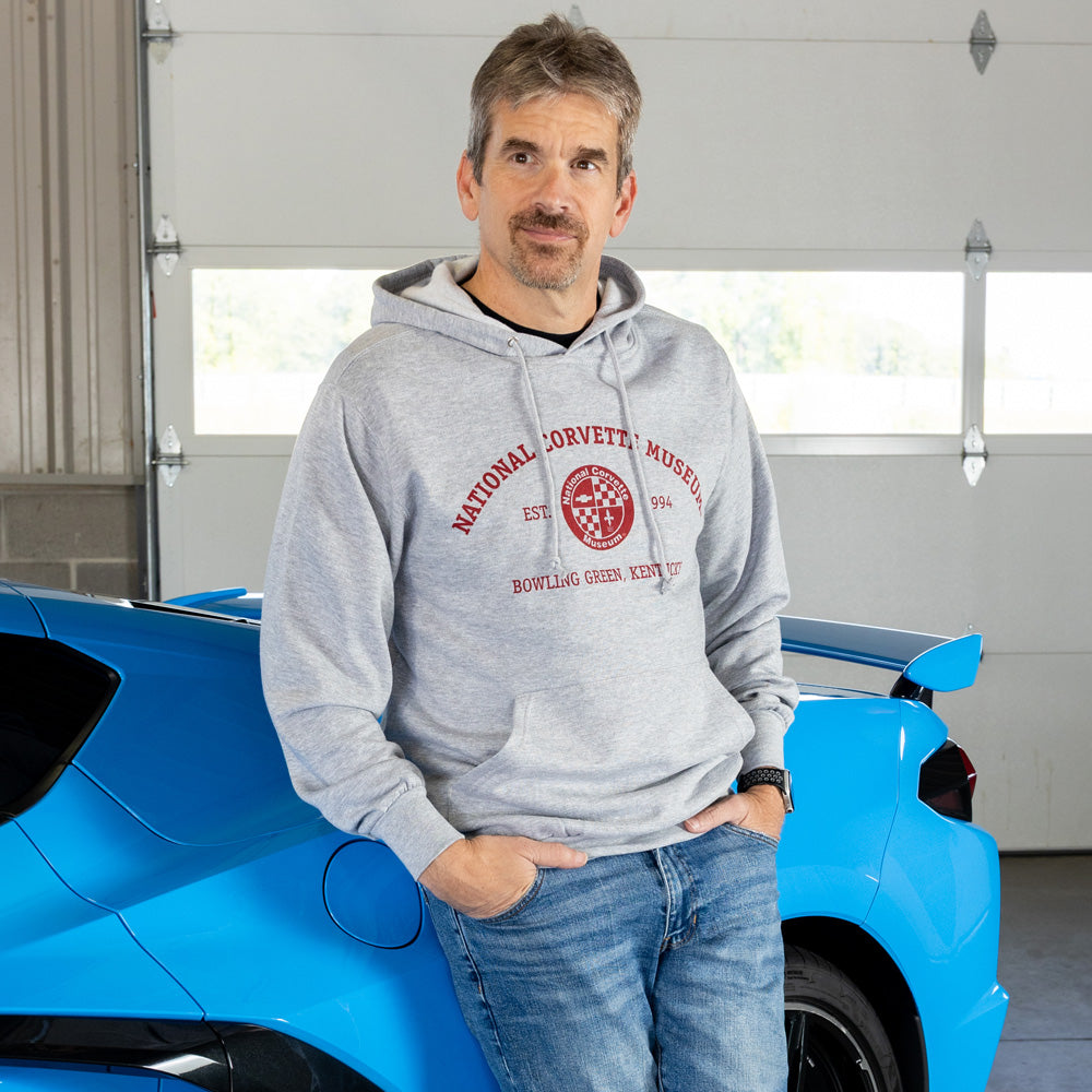 Man wearing a gray hoodie with a National Corvette Museum emblem standing next to a blue Corvette in a garage.