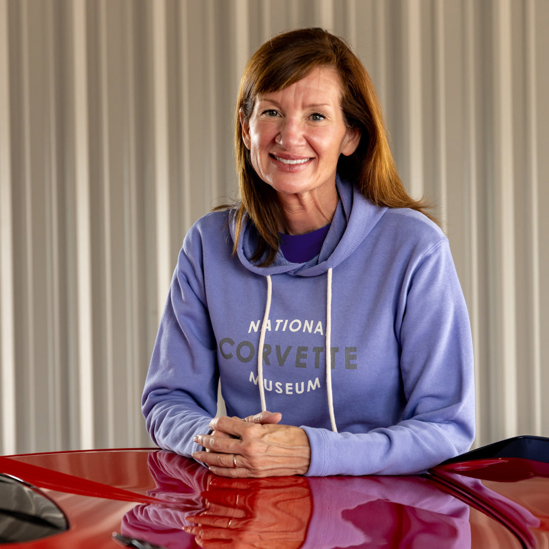Woman wearing a purple hoodie with 'National Corvette Museum' text, sitting in front of a red Corvette.